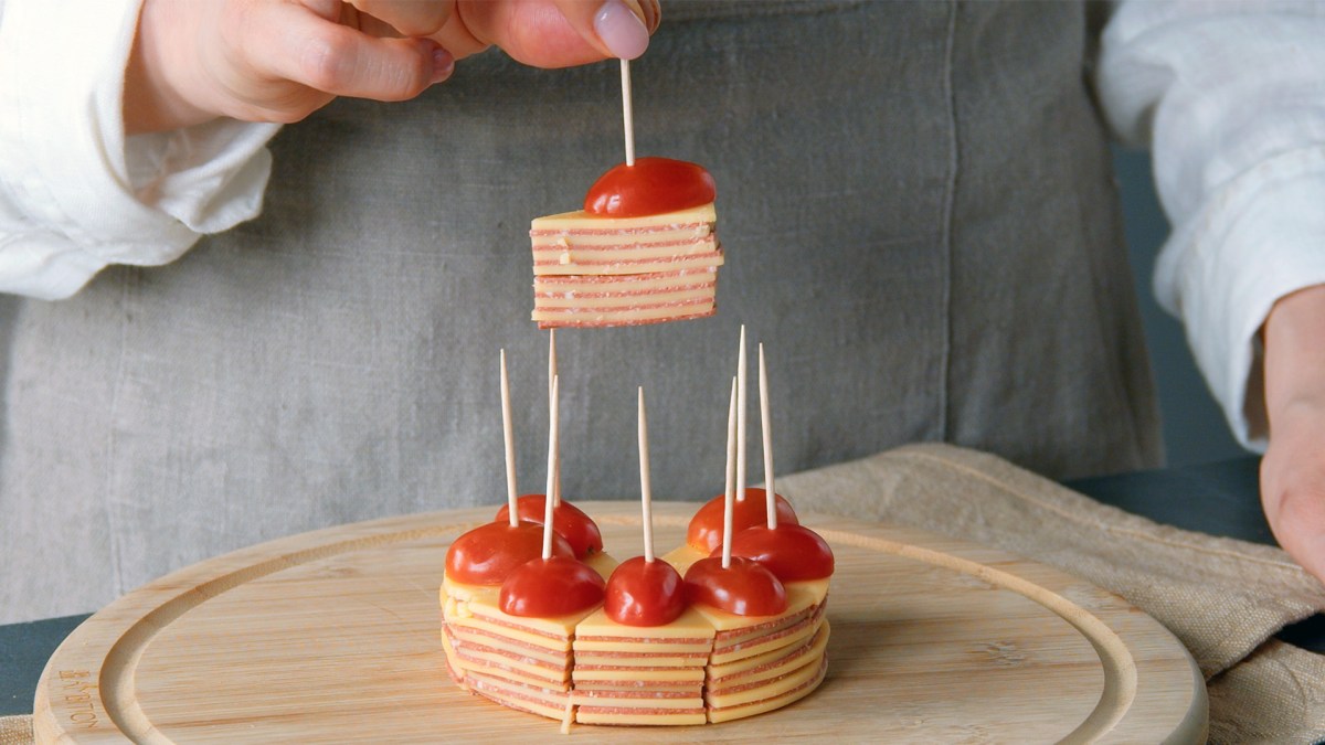 Salami-Käse-Snack mit Tomate auf einem Holzbrett. Die Hand einer Köchin hält ein Stück in die Kamera