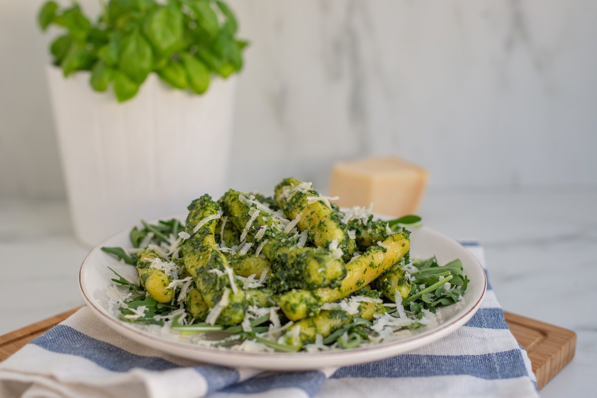 Ein Teller Schupfnudelpfanne mit Spargel und Rucola-Pesto mit Parmesan bestreut steht auf einem blau-weiß-gestreiften Handtuch.