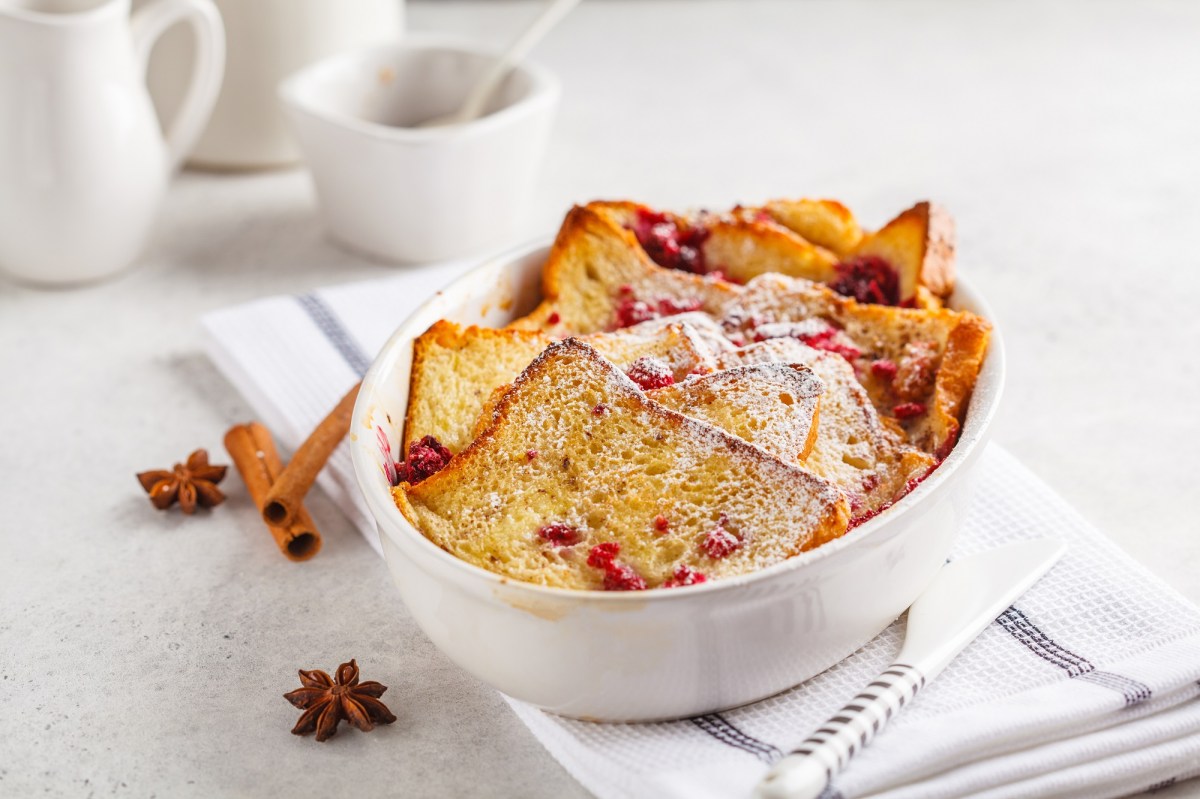 Toastbrotauflauf mit Himbeeren und Puderzucker in weißer Auflaufform, daneben Anissterne und Zimtstangen.