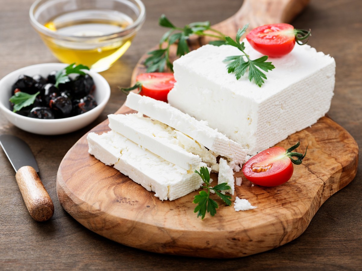 Unterschied Feta, Hirtenkäse und Schafskäse: Ein Stück Käse liegt auf einem Holzbrett. Garniert ist es mit TOmaten und Oliven.