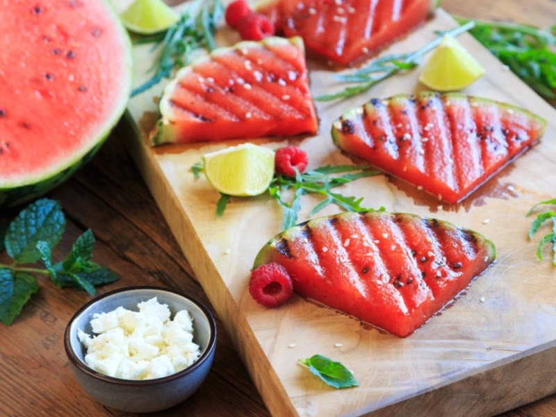 Wassermelonenstücke vom Grill auf einem Holzbrett. Daneben steht eine Schale mit Feta.