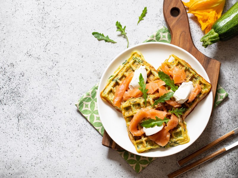 Zucchiniwaffeln mit Lachs auf einem weißen Teller, der auf einem Holzbrett steht.