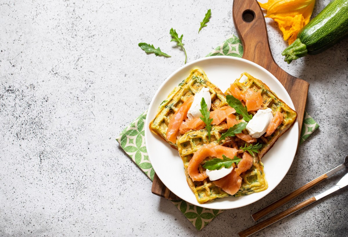 Zucchiniwaffeln mit Lachs auf einem weißen Teller, der auf einem Holzbrett steht.