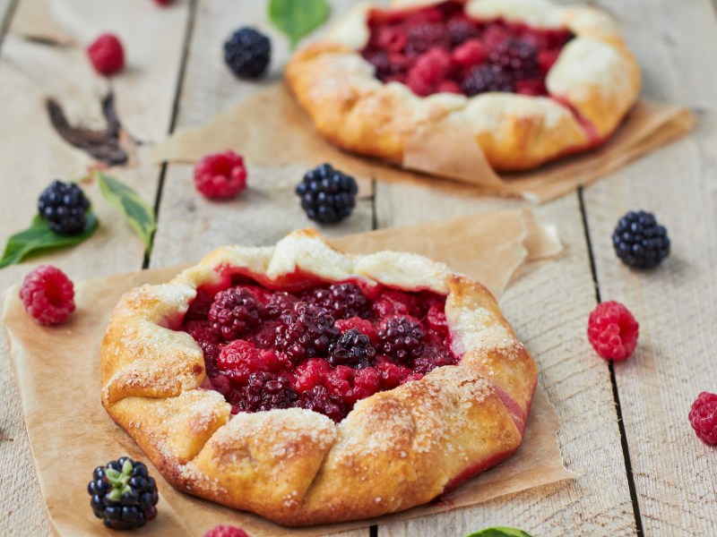 Zwei Beeren-Galettes auf einem Holztisch. Drum herum liegen frische Beeren.