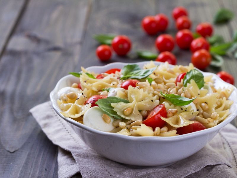 Eine weiße Schüssel Caprese-Nudelsalat mit Tomaten, Mozzarella, Basilikum und Pinienkernen. Im Hintergrund ganze Tomaten am Strauch.