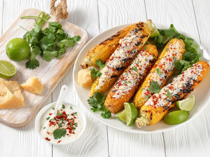 Elote - mexikanischer Streetfood-Mais auf einem weißen Teller. Daneben Limetten, Koriander und Käse.