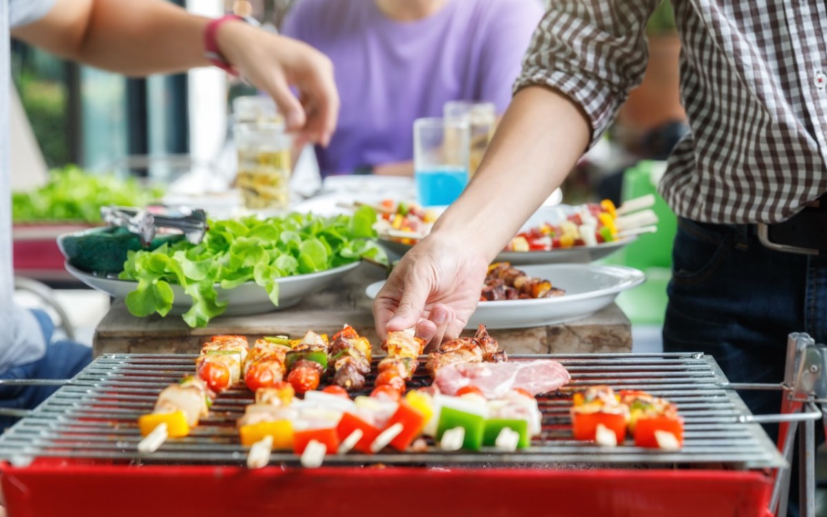 Was zu einem Grillabend dazu gehört: ein Grill mit Spießen im Vordergrund. Dahinter Beialgen und Menschen.