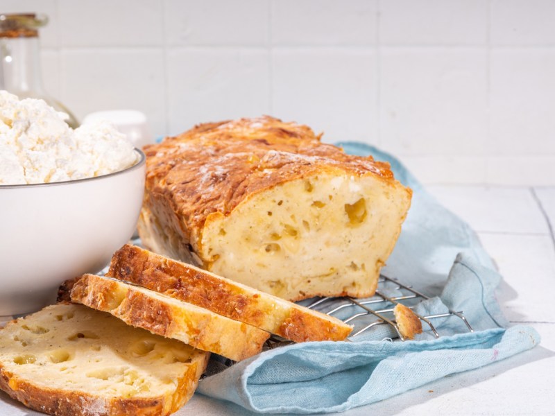 Angeschnittenes Hüttenkäse-Brot auf einer blauen Serviette. Daneben steht eine Schüssel mit Hüttenkäse.