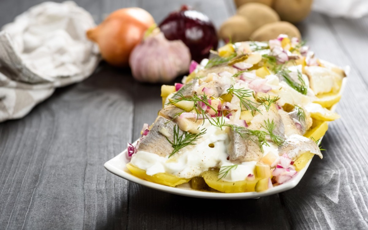 Ein Teller mit Kartoffelsalat mit Hering. Im Hintergrund liegen frische Zutaten.