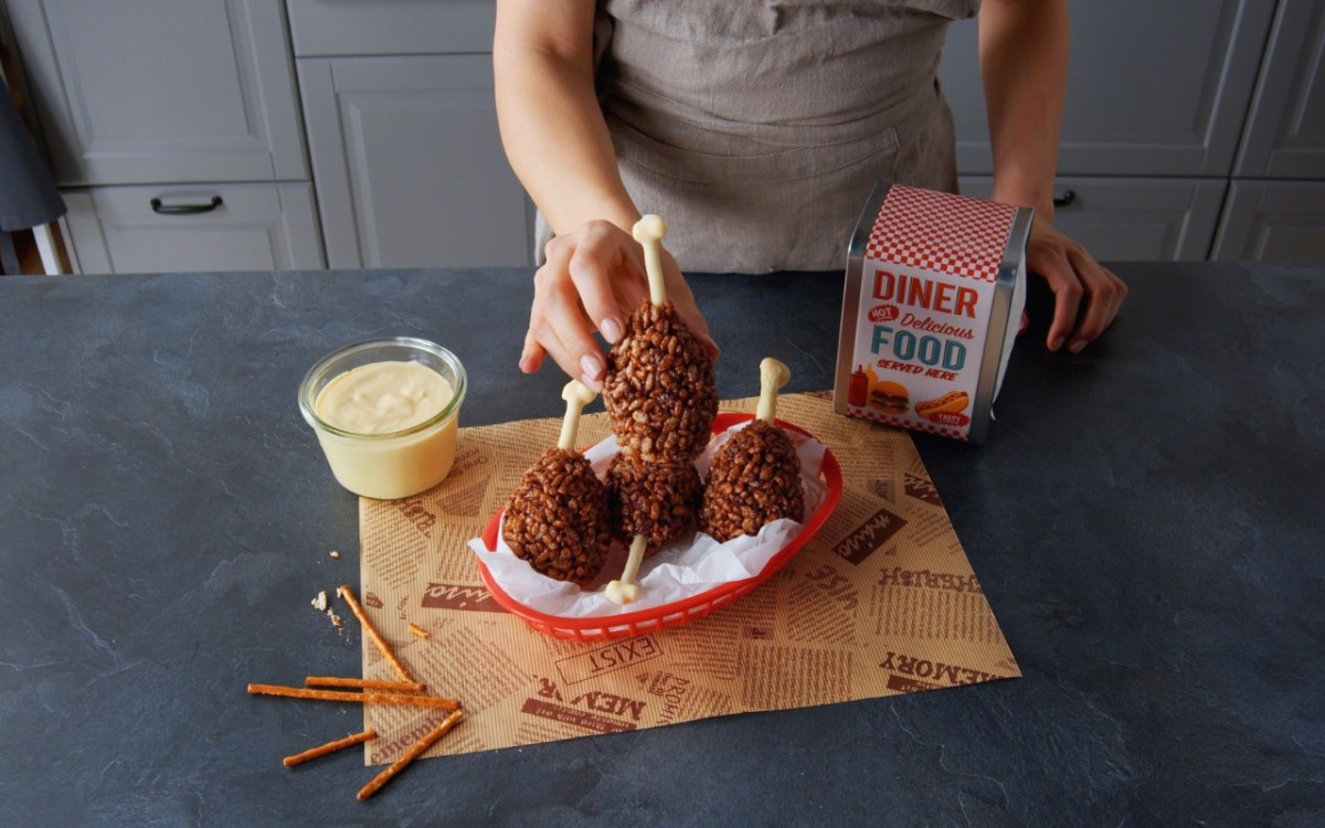 Eine Knusperkeule aus Reiscrispies wird von einer Hand gehalten. Weitere Keulen sind im Hintergrund zu sehen.