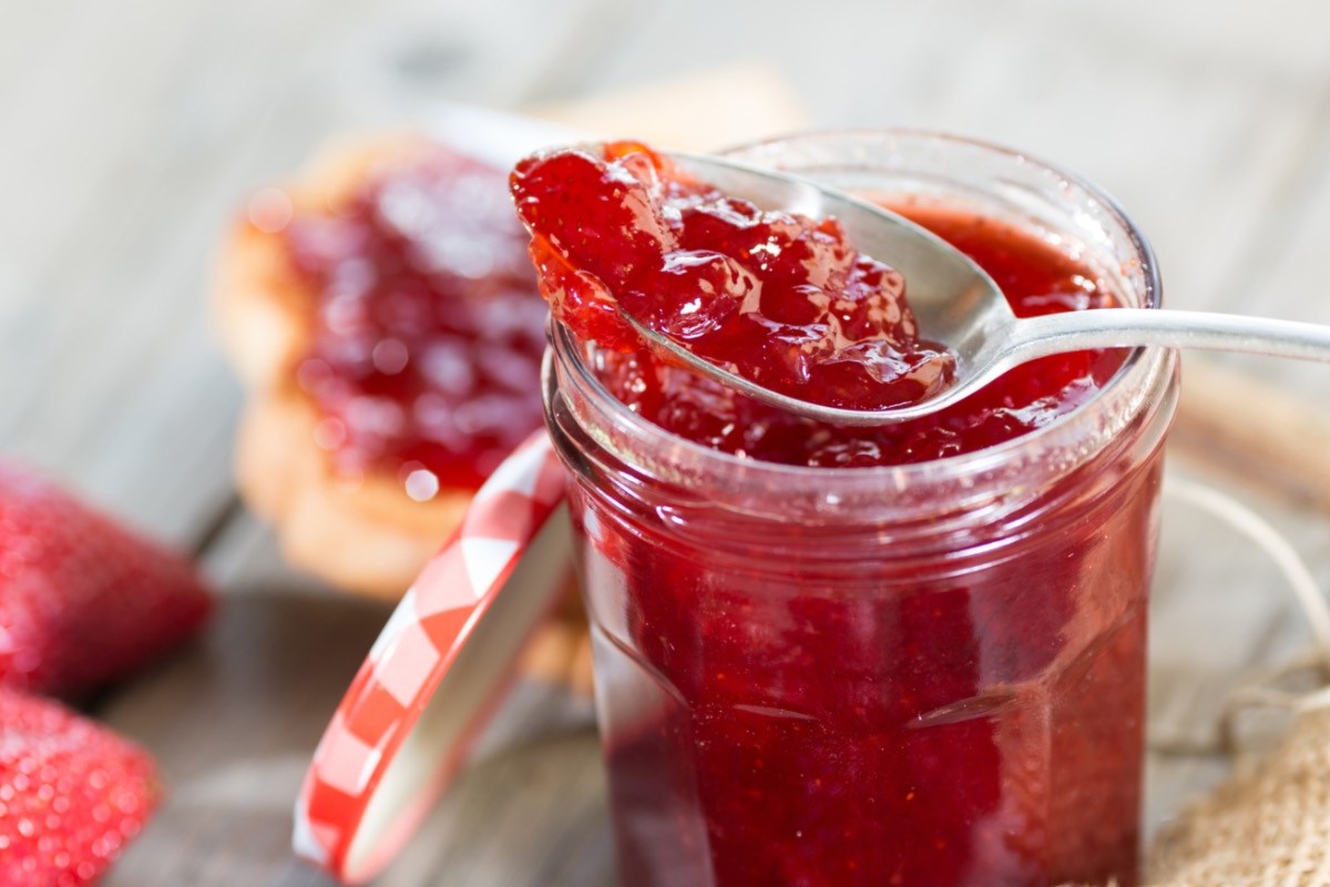 Marmelade einfrieren: ein Glas Erdbeermarmelade mit einem Löffel darin. Im Hintergrund Brötchen mit Marmelade darauf.