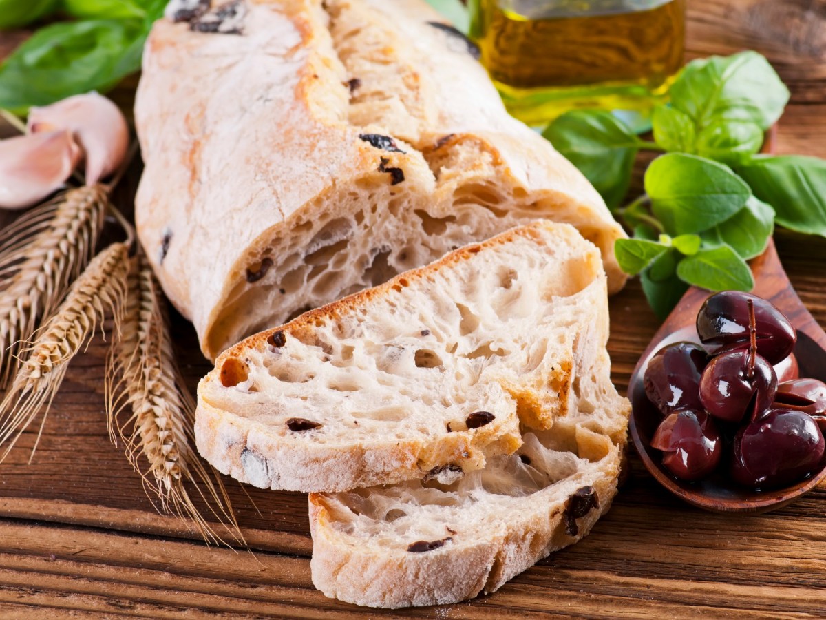 Oliven-Ciabatta auf einem Holzbrett. Es wurde bereits angeschnitten und daneben liegen einige Oliven.