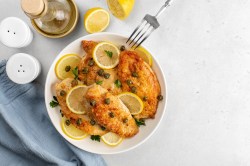 Piccata mit Hähnchen auf einem weißen Teler. Daneben liegt Zitrone und steht eine Flasche Olivenöl.