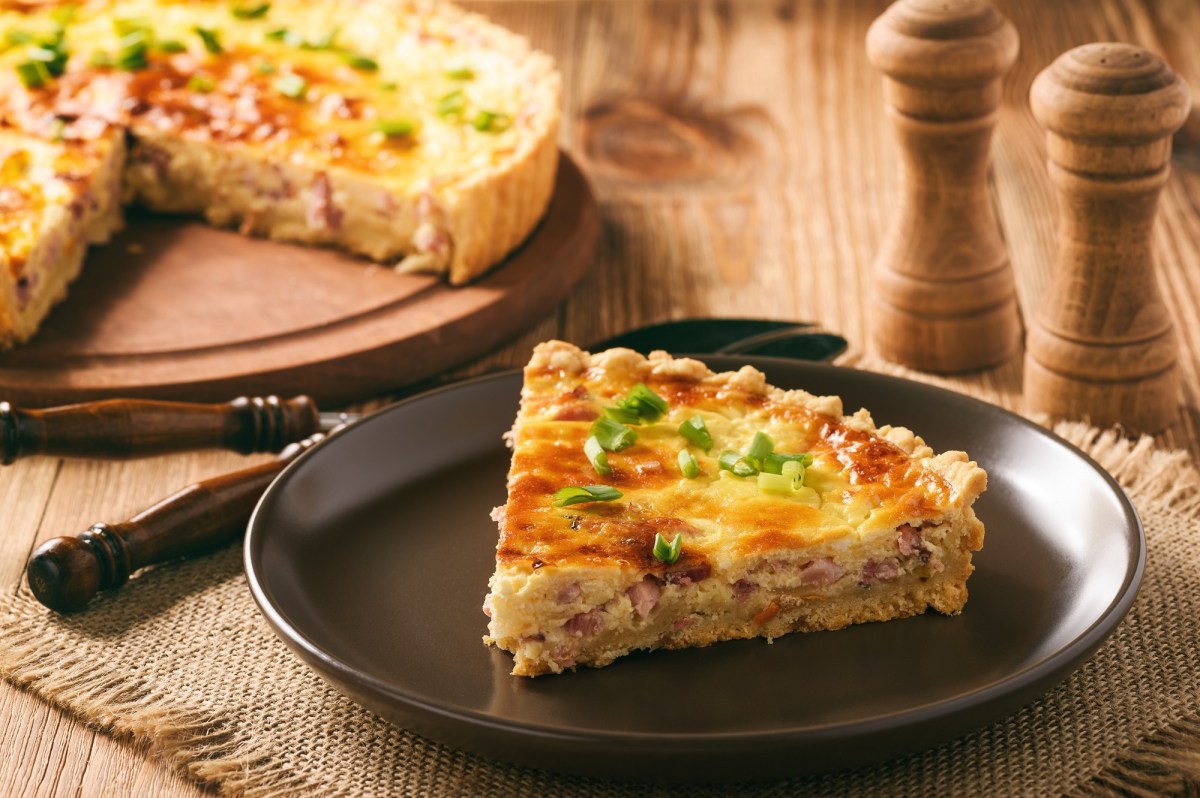 Ein Stücke Quiche Lorraine auf einem dunklen Teller, bestreut mir Frühlingszwiebeln. Im Hintergrund der Rest der Quiche