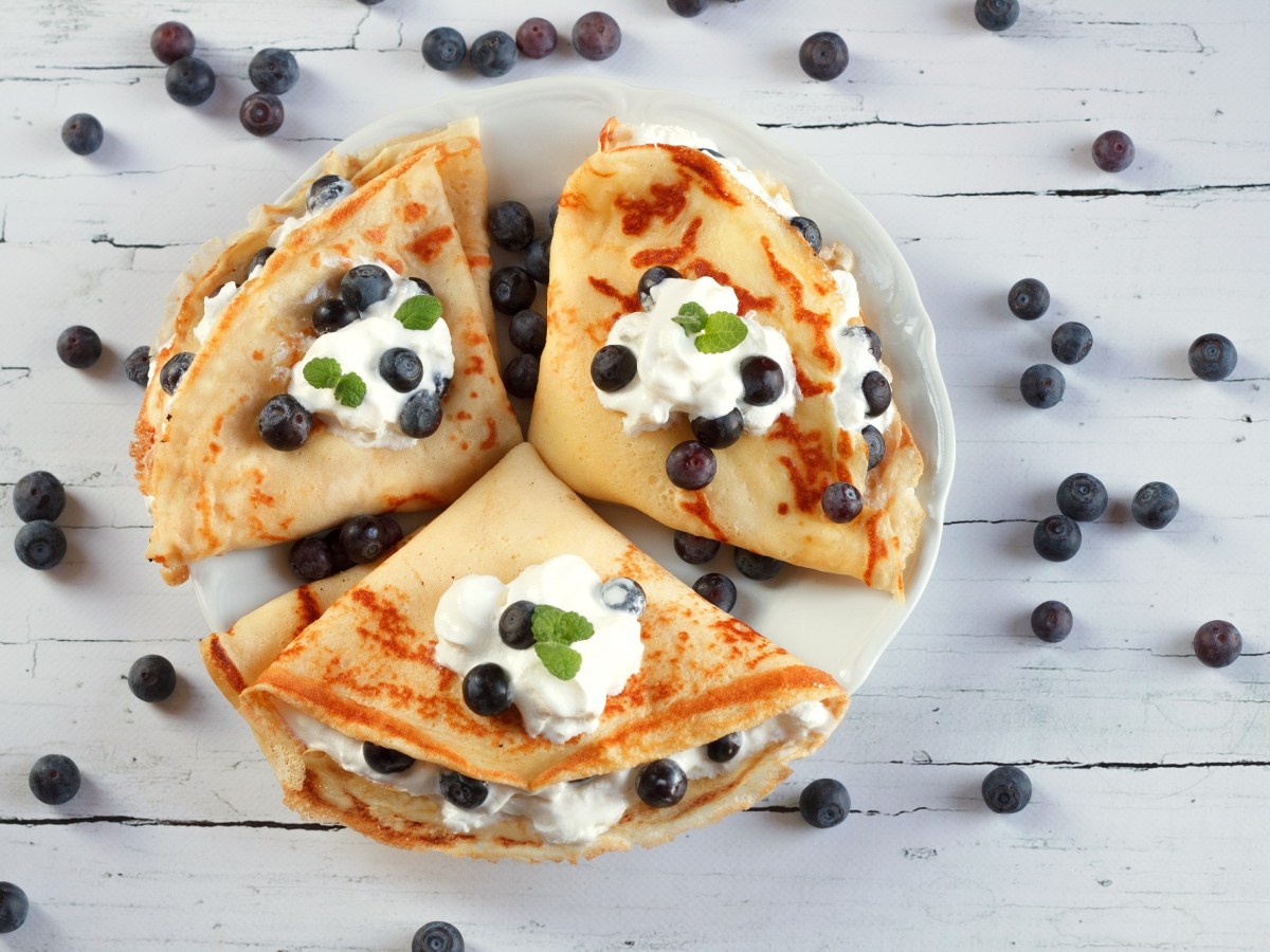 Drei Schmand-Pfannkuchen auf einem weißen Teller. Darauf und daneben liegen frische Heidelbeeren.