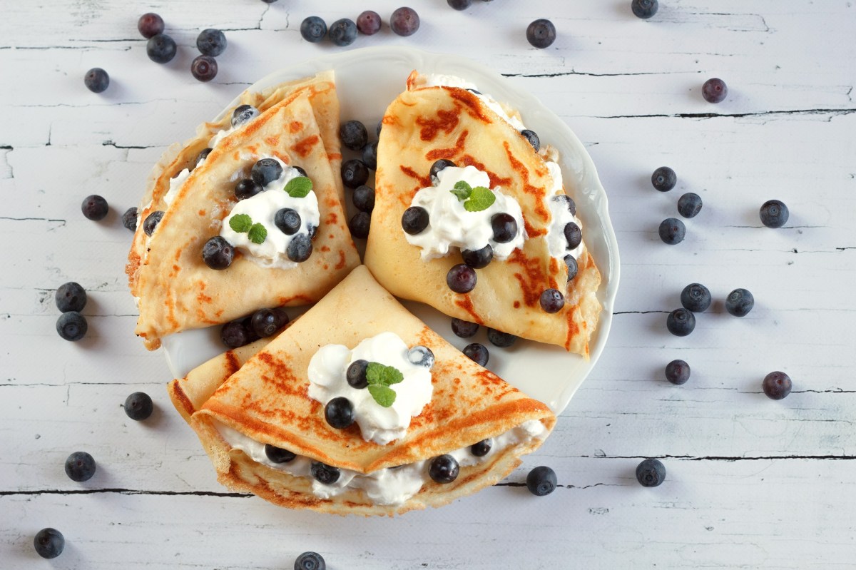 Drei Schmand-Pfannkuchen auf einem weißen Teller. Darauf und daneben liegen frische Heidelbeeren.