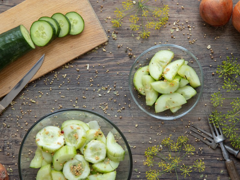 Glas Schüttelgurken mit Gurke auf Holzbrett, Zwiebeln Dill und Senfkörnern.