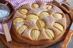 Versunkener Apfelkuchen mit Zimtäpfeln bestreut mit Puderzucker, daneben ein Sieb. Nahaufnahme.