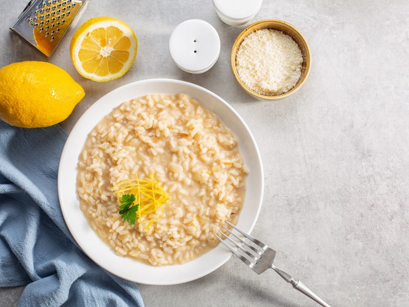 Teller mit Zitronen-Risotto auf hellem Tisch, aufgeschnittene Zitrone, Gabel und Schale mit Parmesan daneben.