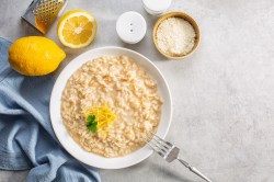 Teller mit Zitronen-Risotto auf hellem Tisch, aufgeschnittene Zitrone, Gabel und Schale mit Parmesan daneben.