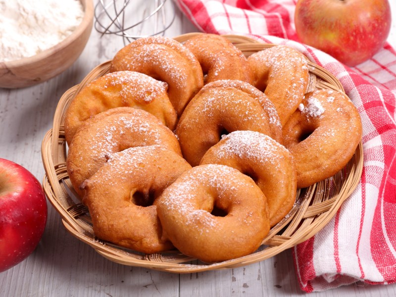 Apfel-Beignets, mit Puderzucker bestreut, in einem Korb. Daneben liegen Äpfel.