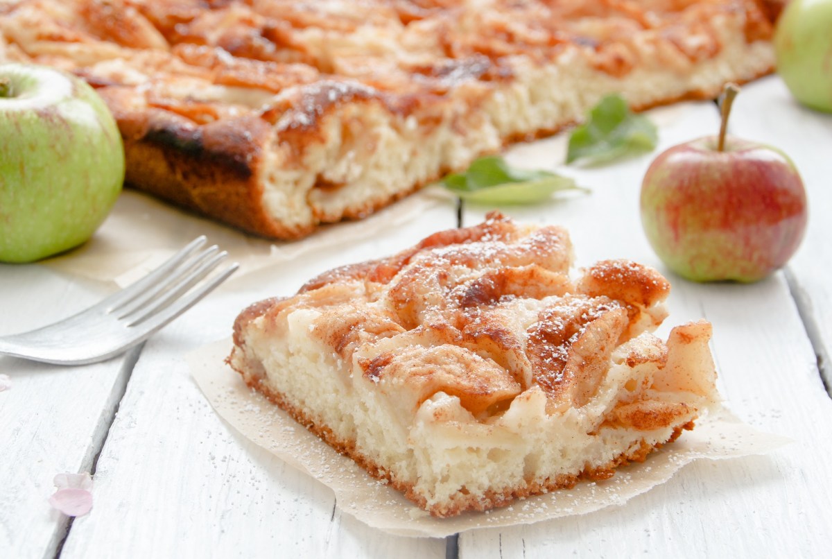 Blechkuchen-Rezepte: ein Stück Apfelkuchen vom Blech auf einem Stück Backpapier, im Hintergrund der Kuchen und 2 Äpfel.