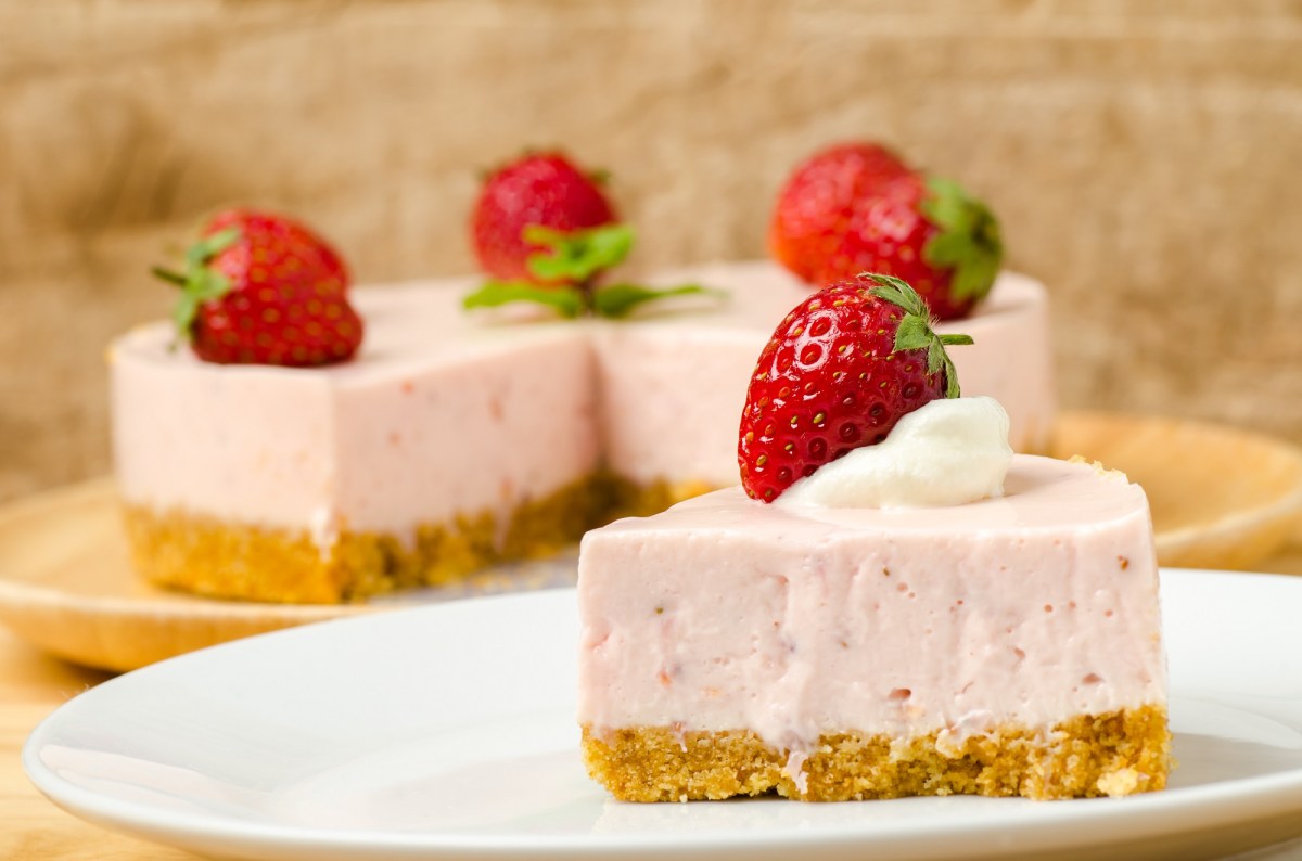 Ein Stück Erdbeermousse-Kuchen, garniert mit einer Erdbeere, auf einem Teller, im Hintergrund der angeschnittene Kuchen.