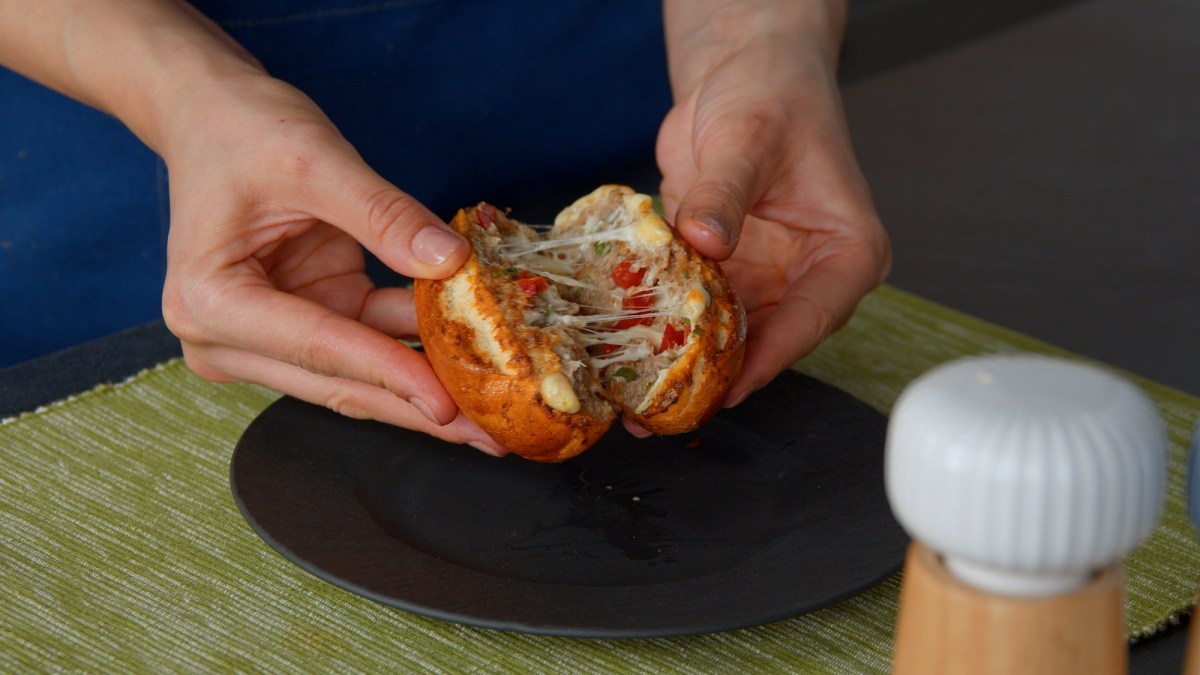 Eine Frau teilt ein gefülltes Hackfleisch-Käsebrötchen, man sieht nur ihre Hände, Nahaufnahme.