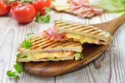 Zwei gegrillte Käse-Schinken-Sandwiches auf einem Brett, im Hintergrund frische Tomaten.