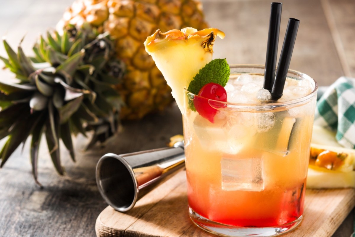 Ein Glas Mai Tai mit zwei Strohhalmen. Dahinter liegen Cocktailzubehör und eine Ananas.