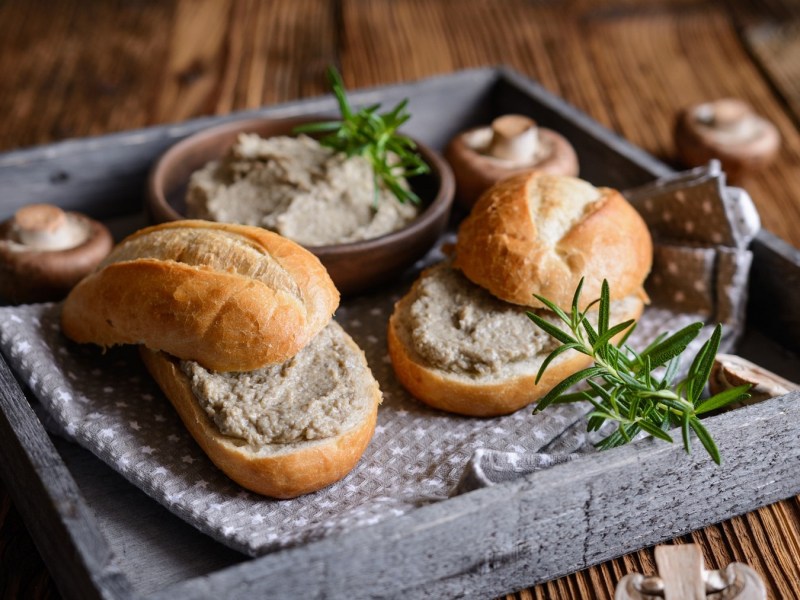 Zwei Brötchen mit Brotaufstrich mit Champignons und Frischkäse auf einem Tablett, daneben Pilze.