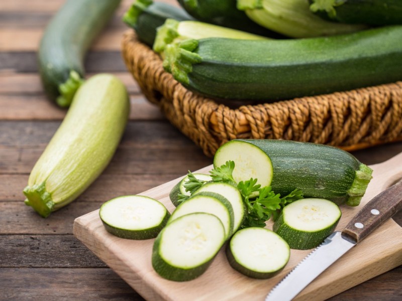 Aufgeschnittene Zucchini auf einem Brett. Im Hintergrund Zucchini in einem Korb.