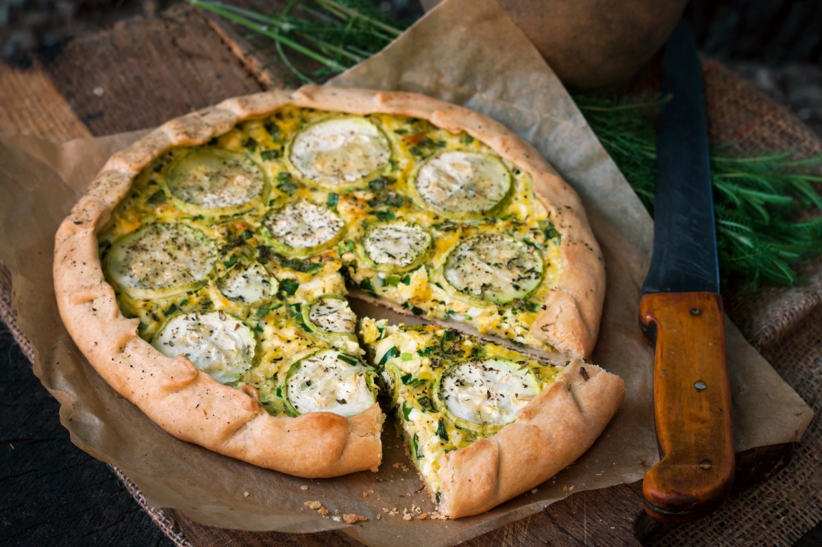 Zucchini-Tarte mit Ziegenkäse auf einem Backpapier, das auf einem Holzbrett liegt. Ein Stück wurde bereits abgeschnitten.