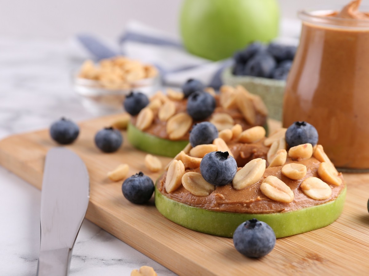 Apfel-Erdnussbutter-Bites auf einem Holzbrett. Sie sind mit Blaubeeren und Erdnüssen garniert.