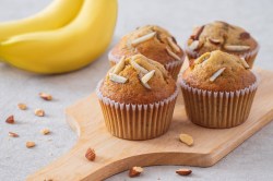 4 Bananenmuffins ohne Zucker auf einem Brett, garniert mit Mandelstiften, dahinter Bananen.