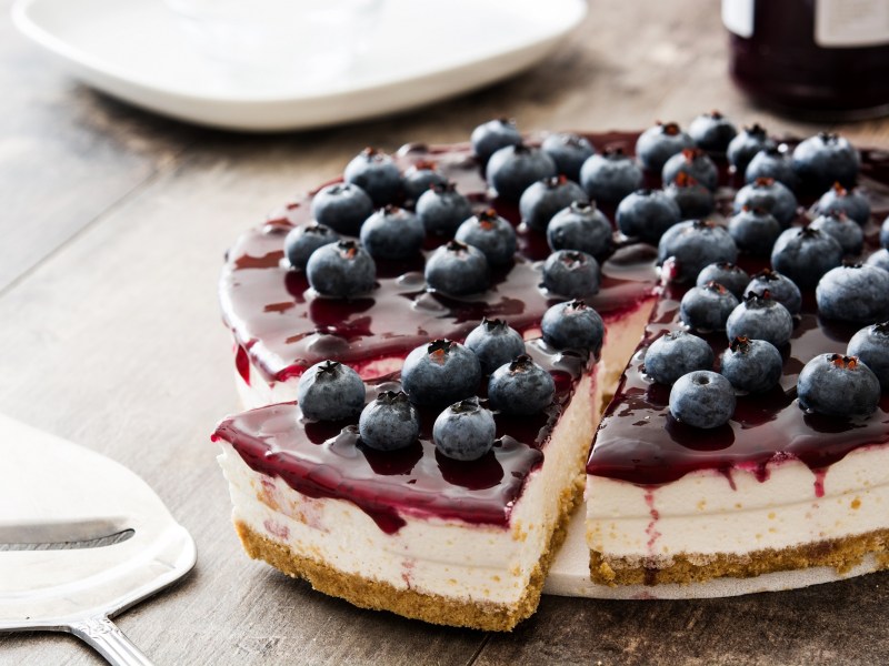 Ein Heidelbeer-Quarkkuchen, von dem ein Stück abgeschnitten wird, auf einem weißen Teller.