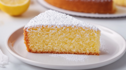 Ein Stück italienischer Ricotta-Zitronenkuchen mit Puderzucker auf einem Teller, dahinter unscharf eine halbe Zitrone und der Kuchen.