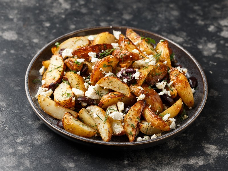 Ein tiefer Teller Kartoffel-Wedges mit Feta und Oliven auf dunklem Marmoruntergrund.