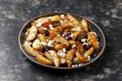 Ein tiefer Teller Kartoffel-Wedges mit Feta und Oliven auf dunklem Marmoruntergrund.