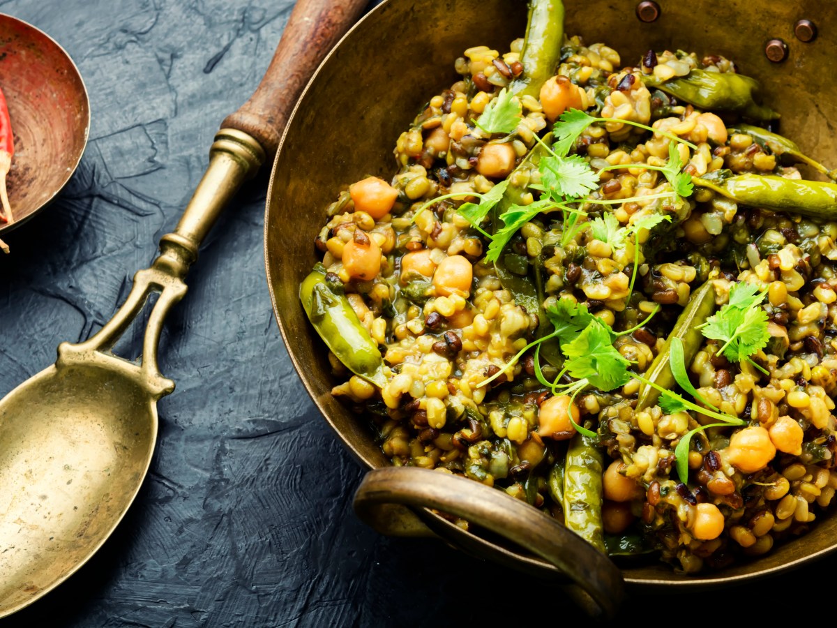 Kitchari, indischer Kokosreis mit Bohnen und Kichererbsen, in einer Blechschüssel, daneben ein Löffel. Draufsicht.