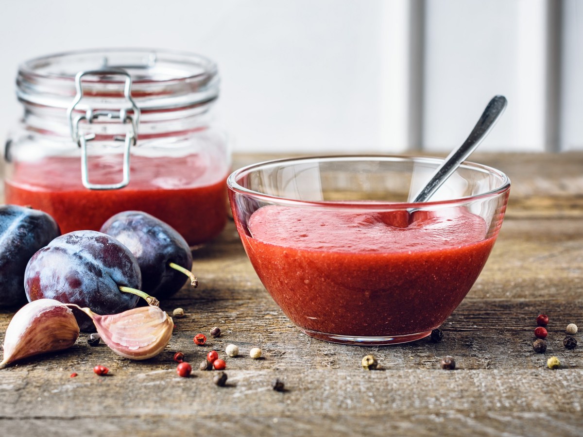 Pflaumenketchup in einer Schale. Daneben liegen Pflaumen und Knoblauchzehen. Im Hintergrund steht ein Glas mit mehr Ketchup.