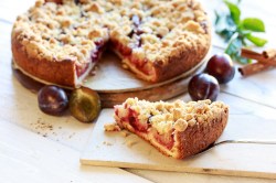 angeschnittener Pflaumenkuchen mit Zimtstreuseln, ein Kuchenstück liegt auf einem Brett davor.