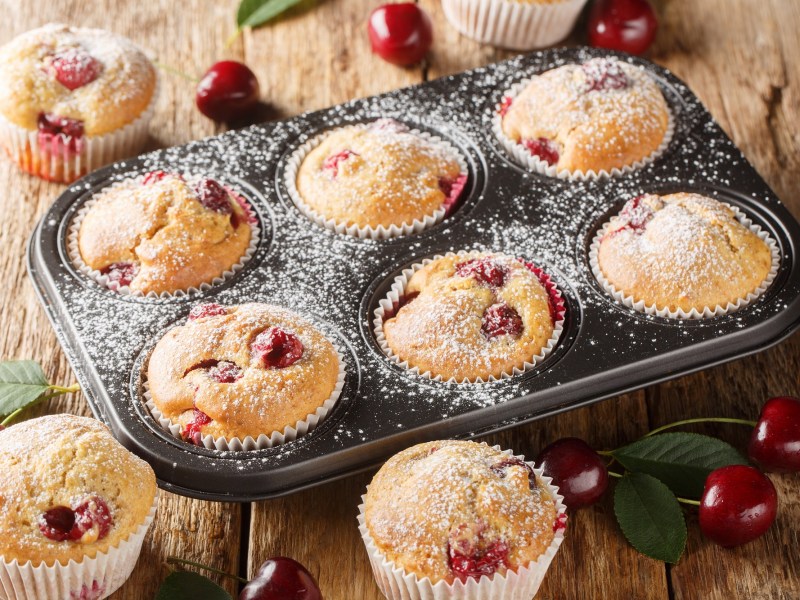 Ein 6-er Muffinblech mit Quark-Muffins mit Kirschen, daneben frische Kirschen und mehr Muffins.