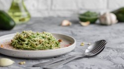 Ein Teller Zucchini-Avocado-Pasta garniert mit Pinienkernen und Parmesan, daneben ein Löffel.