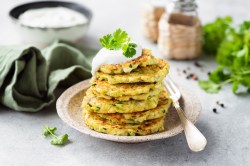 Ein Stapel Zucchini-Pancakes mit Hüttenkäse auf einem weißen Teller. Daneben liegt eine Gabel. Die Pancakes sind mit Joghurt und Petersilie garniert.