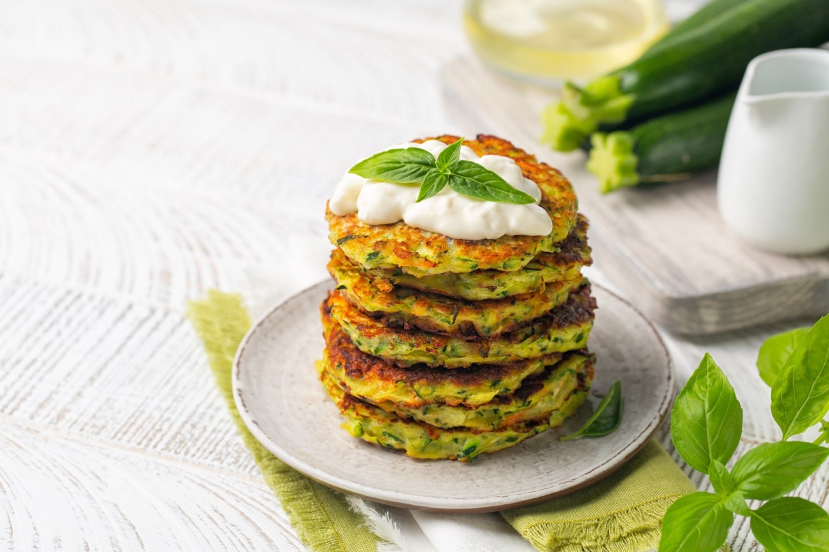 Ein Stapel Zucchini-Parmesan-Puffer auf einem Teller, garniert mit einem Klecks Schmand und Basilikum.