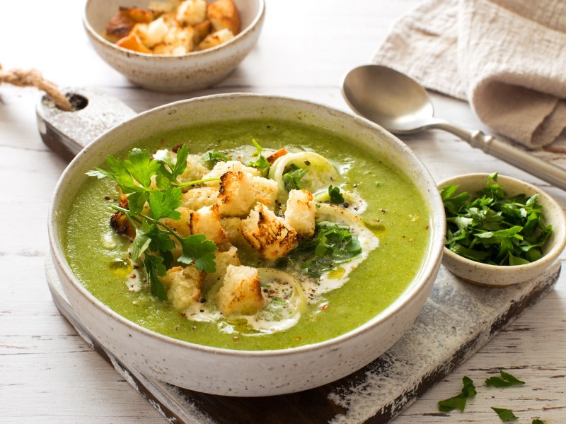 Eine Schüssel Zucchini-Spinat-Suppe mit Croûtons, Lauch, Schmand und Kräutern.