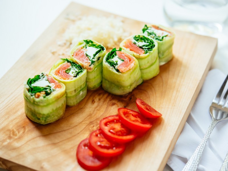 6 Gurken-Röllchen mit Lachs und Feta auf einem Brett, daneben in Scheiben geschnittene Tomaten.