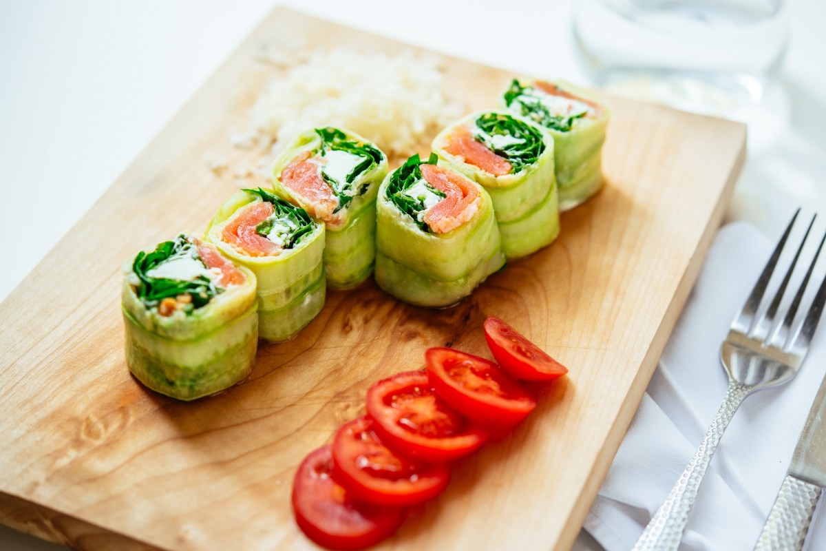 6 Gurken-Röllchen mit Lachs und Feta auf einem Brett, daneben in Scheiben geschnittene Tomaten.