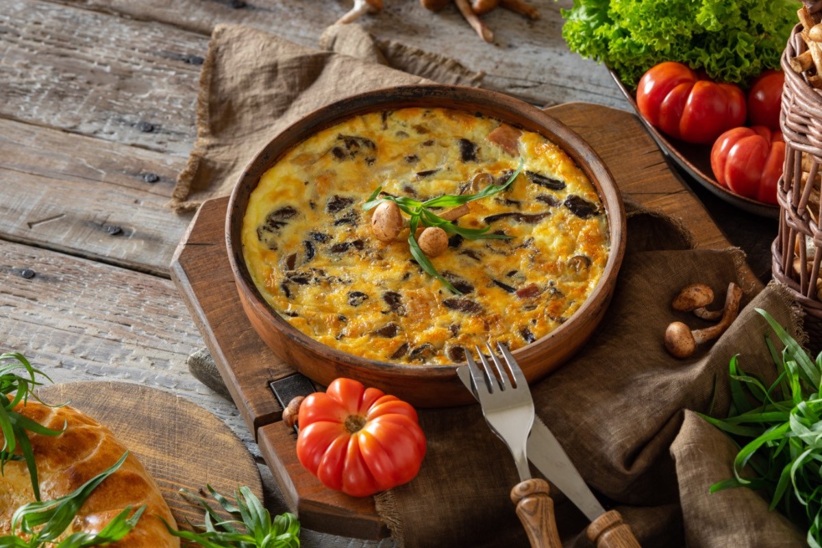 Eine Kartoffel-Frittata mit Pilzen in einer Pfanne. Drumherum liegen frische Zutaten.